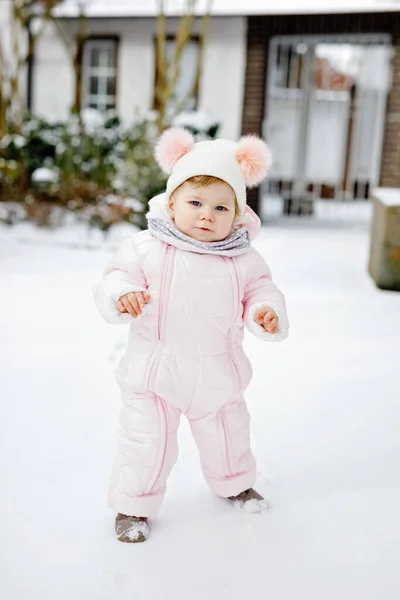 Lycklig liten flicka gör första stegen utomhus på vintern genom snö. Söta småbarn som lär sig gå. Barn har kul på kall snöig dag. Bär varma baby rosa kläder snödräkt och bubblor hatt. — Stockfoto