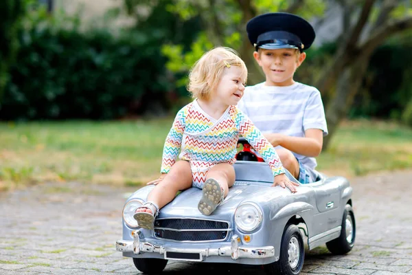 Duas crianças felizes brincando com o carro de brinquedo velho grande no jardim de verão, ao ar livre. Rapaz carro de condução com a menina da criança lá dentro. Miúdos risonhos e sorridentes. Família, infância, conceito de estilo de vida — Fotografia de Stock