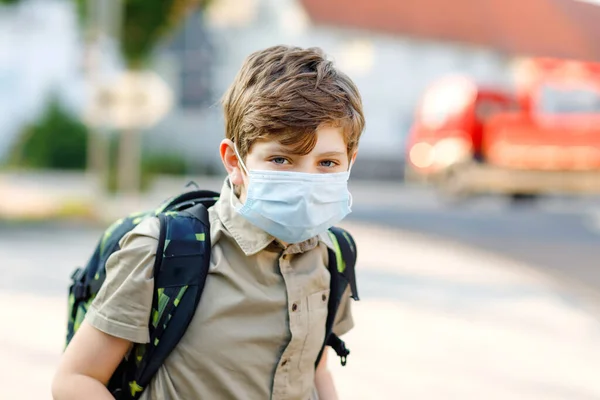 Glücklicher Junge mit Brille und medizinischer Maske wegen Coronavirus-Pandemie. Schulkind mit Schulranzen wartet am sonnigen Tag auf dem Schulweg auf den Bus. Gesundes Kind auf der Straße. — Stockfoto