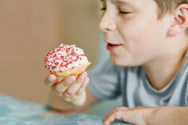 Buon ragazzo della scuola preadolescente mangiare ciambella dolce al coperto. Bambino biondo in cucina domestica o nella mensa scolastica Bambino carino e cibo zuccherato malsano. — Foto Stock