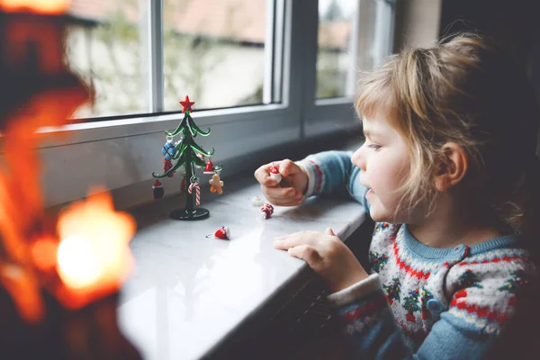 Dziewczynka Litte Maluch siedzi przy oknie i dekoracji małej choinki szkła z małych zabawek xmas. Szczęśliwe zdrowe dziecko świętować rodzinne tradycyjne wakacje. Urocze dziecko.. — Zdjęcie stockowe