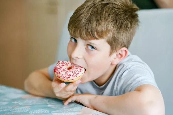 Happy preteen school boy eating sweet donut indoor. Blond child in domestic kitchen or in school canteen Cute kid and unhealthy sugared food. — Stock Photo, Image