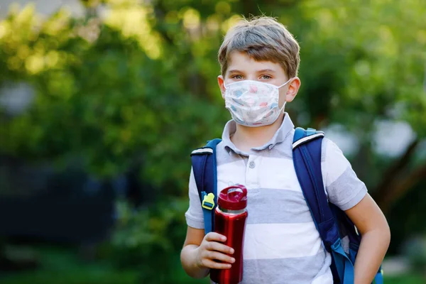 Niño feliz, máscara médica, botella de agua y mochila o mochila. Un colegial de camino a la escuela. Un niño sano al aire libre. Regreso a la escuela después de la cuarentena del bloqueo de la enfermedad pandémica de corona — Foto de Stock