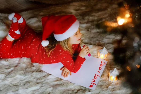 Pequeña niña linda en la cama bajo el árbol de Navidad, escribir carta lista de deseos a Santa Claus en casa, en el interior. Fiesta cristiana tradicional. Niño feliz esperando regalos en Navidad. — Foto de Stock