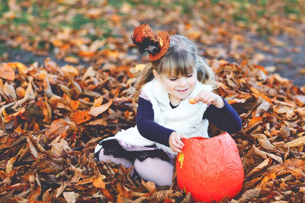 Little toddler girl dressed as a witch trick or treating on Halloween. Happy child outdoors, with orange funny hat and pumpkin bag for sweet haunt. Family festival season in october. Outdoor activity — Stock Photo, Image