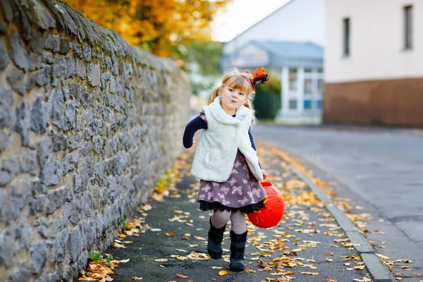 魔女のトリックやハロウィンの扱いに身を包んだ小さな幼児の女の子。幸せな子供は屋外で、オレンジ面白い帽子とカボチャの袋で甘い出没します。10月のファミリーフェスティバルシーズン。野外活動 — ストック写真