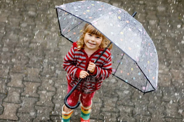 雨の日に大きな傘で遊んでいる小さな幼児の女の子。雨の中を走る幸せな正の子、水たまり。雨の服やゴムブーツを持つ就学前の子供。悪天候の日の子供の活動. — ストック写真