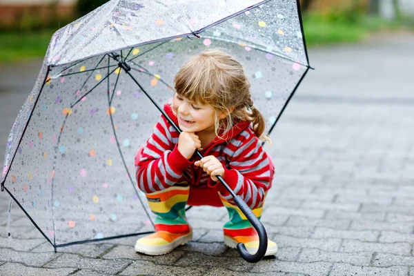 Little toddler girl playing with big umbrella on rainy day. Happy positive child running through rain, puddles. Preschool kid with rain clothes and rubber boots. Children activity on bad weather day. — Stock Photo, Image