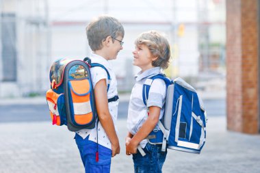 Sırt çantası ya da çantası olan iki küçük çocuk. Okulun yolu üzerindeki okul çocukları. Sağlıklı sevimli çocuklar, erkek kardeşler ve en iyi arkadaşlar sokakta evden ayrılıyor. Okula dönelim. Mutlu kardeşler..