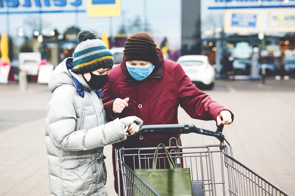 Oude oudere vrouw en kind jongen met medisch masker als gevolg van pandemische coronavirus ziekte. Kinderhulp oma met winkelen in de supermarkt. Kleinzoon en oma duwen kar trolley samen, buiten — Stockfoto