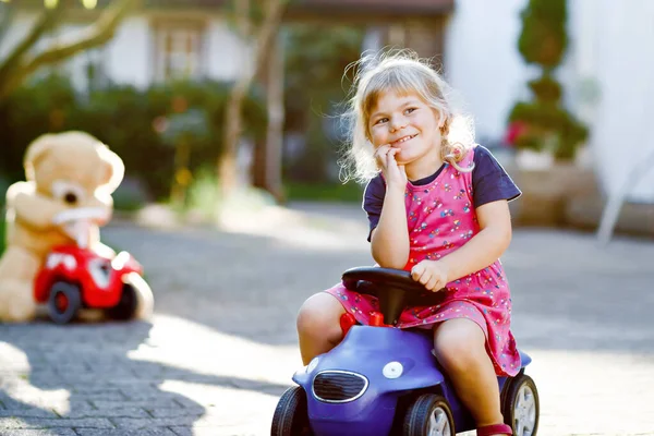 Pequeña niña adorable conduciendo un coche de juguete y divirtiéndose jugando con un oso de peluche, al aire libre. Precioso niño feliz y saludable disfrutando de un cálido día de verano. Sonriente niño impresionante en gaden — Foto de Stock