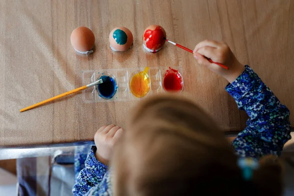 Closeup de menina pequena colorir ovos para a Páscoa. Close-up de criança olhando surpreso com ovos coloridos coloridos, celebrando férias com a família. De cima, rosto não reconhecido. — Fotografia de Stock