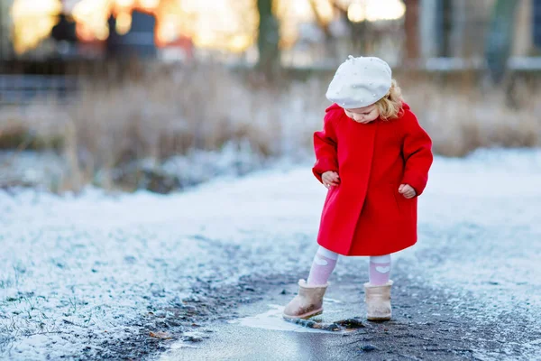 Utomhus vintern porträtt av små söta småbarn flicka i röd rock och vitt mode hatt barret. Friska glada barn promenader i parken på kall dag med snö och snöfall. Snygga kläder för barn. — Stockfoto