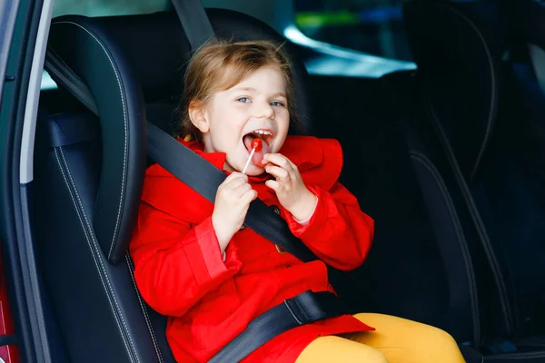 Little toddler girl sitting in car seat, holding sweet lollipop against nausea and sickness and looking out of the window on nature and traffic. Cute kid traveling by car. Child safety on the road. — Stock Photo, Image