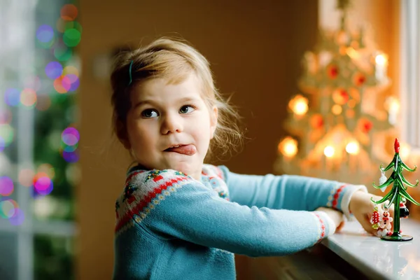 Menina pequena criança sentada à janela e decorando pequena árvore de Natal de vidro com minúsculos brinquedos xmas. A criança sã feliz celebra férias tradicionais de família. Adorável bebê. — Fotografia de Stock