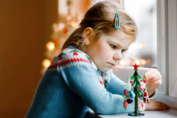 Маленькая девочка сидит у окна и украшает маленькую елку маленькими рождественскими игрушками. Счастливый здоровый ребенок отмечает традиционный семейный праздник. Очаровательный ребенок. — стоковое фото