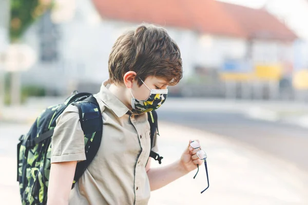 Niño feliz con gafas y máscara médica debido a la pandemia covid virus corona. Colegial con mochila esperando el autobús en el camino a la escuela en el día soleado. Niño sano al aire libre en la calle. — Foto de Stock