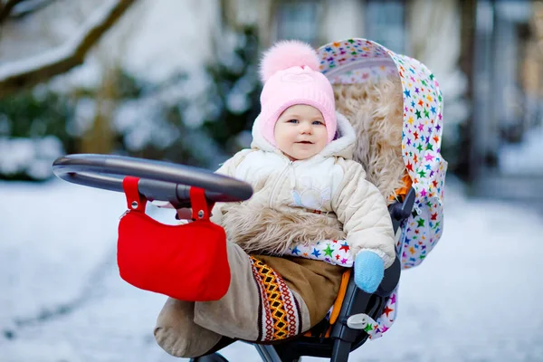 Bonito pequena menina bonita sentada no carrinho de bebê ou carrinho no dia frio de inverno nevado. Criança sorridente feliz em roupas quentes, moda casaco de bebê elegante. Bebês primeira neve. — Fotografia de Stock