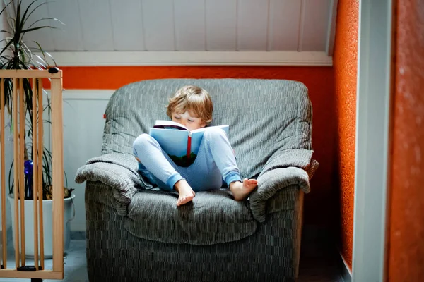 Милый блондин, маленький дошкольник в пижаме, читает книжку для детей в домашней комнате. Взволнованный ребенок читает громко, сидит на старом большом кресле. Школьник, семья, образование — стоковое фото