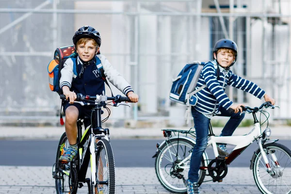 Två skolpojkar i skyddshjälm rider med cykel i staden med ryggsäckar. Glada barn i färgglada kläder cykling på cyklar på väg till skolan. Säkert sätt för barn utomhus till skolan — Stockfoto