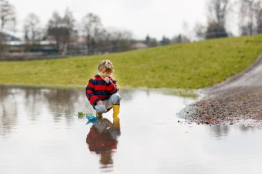 Sarı çizmeli mutlu çocuk baharda ya da sonbahar günü büyük su birikintisinin yanında kağıt gemi teknesiyle oynuyor.