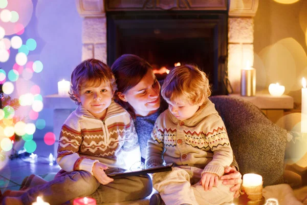 Mladá matka a jeho dvě malé děti sedí u krbu doma na Vánoce — Stock fotografie