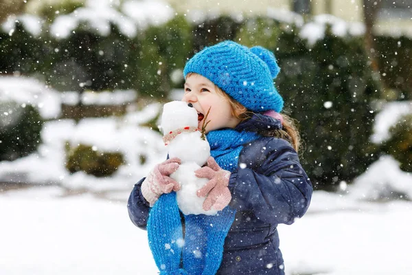 Linda niñita haciendo mini muñeco de nieve y comiendo la nariz de zanahoria. Adorable niño feliz saludable jugando y divirtiéndose con la nieve, al aire libre en el día frío. Ocio activo con niños en invierno —  Fotos de Stock