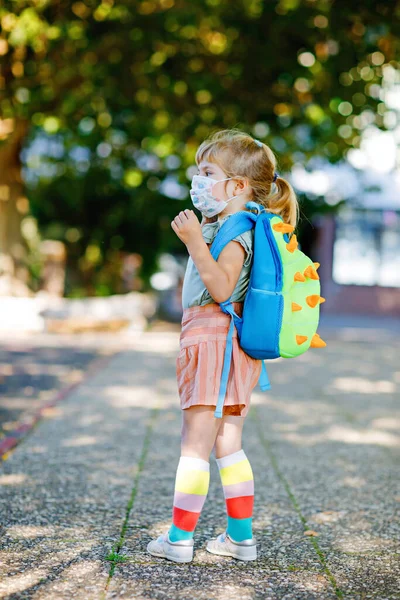 Маленькая девочка-малышка в свой первый день на пути в детскую школу в медицинской маске против коронного ковидного вируса. Здоровый красивый ребенок идет в детский сад и детский сад. Счастливый ребенок с рюкзаком — стоковое фото