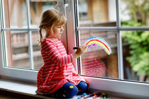 Adoralbe kleine peuter meisje met regenboog geschilderd met kleurrijke venster kleur tijdens pandemische coronavirus quarantaine. Kind schilderen regenbogen en harten over de hele wereld met woorden Laten we allemaal goed zijn. — Stockfoto