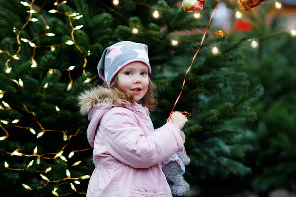 Cute little smiling preschool girl on German Christmas market. Happy child in winter clothes and toy choosing xmas tree on with lights on background. Family, tradition, celebration concept. — Stock Photo, Image