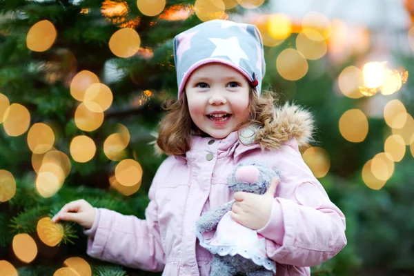 Cute little smiling preschool girl on German Christmas market. Happy child in winter clothes and toy choosing xmas tree on with lights on background. Family, tradition, celebration concept. — Stock Photo, Image
