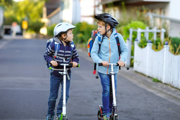 Güneşli bir günde, ellerinde scooterla şehirde gezen kasklı iki okul çocuğu. Renkli giysiler içinde okula bisikletle giden mutlu çocuklar.. — Stok fotoğraf