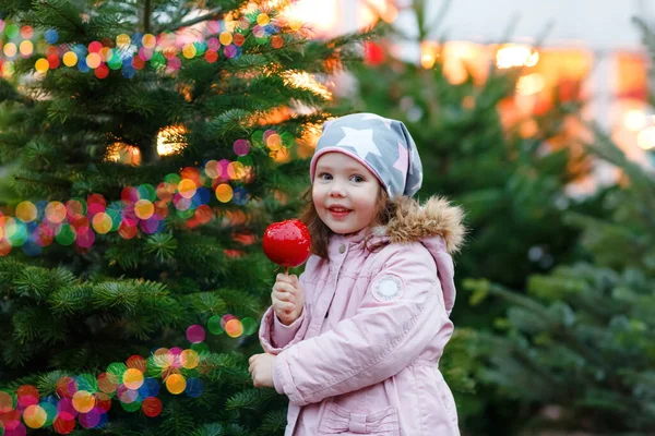독일 크리스마스 마켓에서 귀엽게 미소짓고 있는 유치원 여학생. 겨울 옷을 입은 행복 한 어린이는 배경 조명을 켜 놓고 단맛 이 나는 유약 성분의 사과를 먹는다. 가족, 전통, 축하의 개념 — 스톡 사진