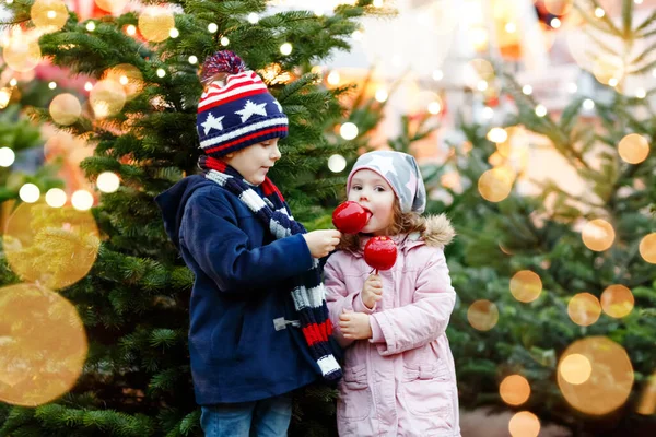 Dwoje uśmiechniętych dzieci, przedszkolak i dziewczynka jedzą słodkie jabłko na niemieckim jarmarku świątecznym. Szczęśliwe rodzeństwo dzieci ze światłami na tle i choinki. Rodzina zabawny brat i siostra. — Zdjęcie stockowe