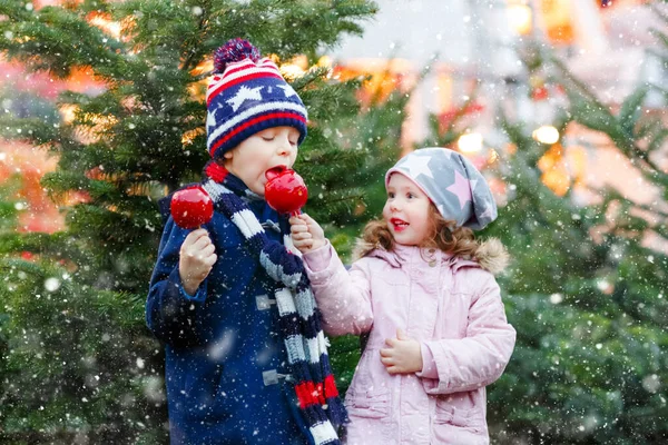 Duas crianças sorridentes, pré-escolar e menina comem maçã açucarada doce no mercado de Natal alemão. Feliz irmãos crianças com luzes no fundo e árvores de xmas. Família engraçado irmão e irmã. — Fotografia de Stock