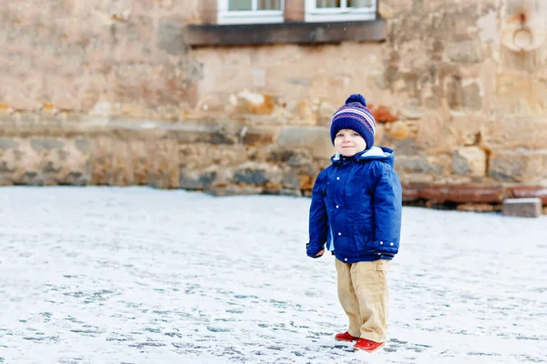 Mały chłopczyk spacerujący po śnieżnym mieście podczas opadów śniegu. Cute szczęśliwy dzieciak w zimie ubrania zabawy na świeżym powietrzu, w zimny dzień ze śniegiem na ulicy. — Zdjęcie stockowe