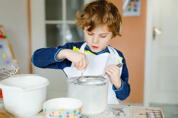 Adorable funny blond little kid boy baking chocolate cake and tasting dough in domestic kitchen, indoors. Happy child having fun with working with mixer, flour, eggs dough at home. Little helper — Stock Photo, Image