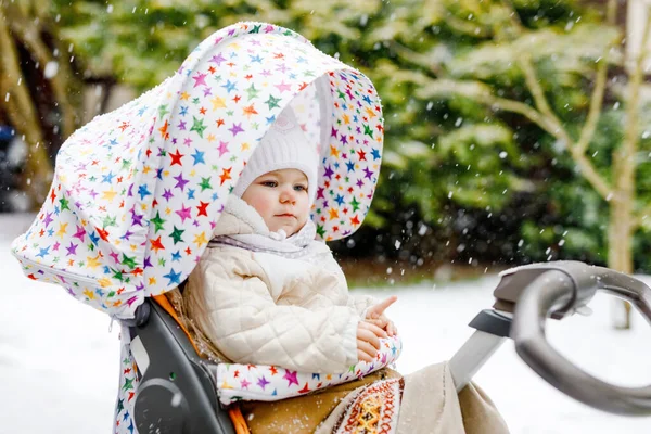 Schattig klein mooi baby meisje zittend in de kinderwagen of kinderwagen op koude besneeuwde winterdag. Gelukkig lachend kind in warme kleren, mode stijlvolle babyjas. Babys eerste sneeuw. Winterwandeling buiten. — Stockfoto