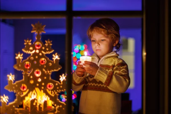 Cute child standing by window at Christmas time and holding cand — Stock Photo, Image