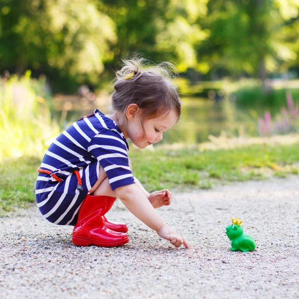 Красивая маленькая девочка в красных ботинках дождя играет с резиновой лягушкой — стоковое фото