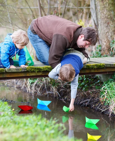 Dos hermanos pequeños y su padre jugando con barcos de papel por un —  Fotos de Stock