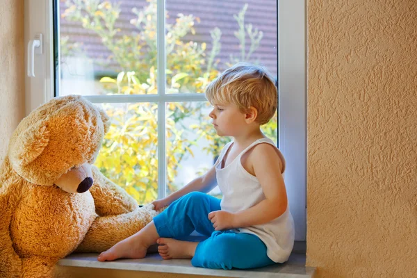 Precioso niño de dos años sentado cerca de la ventana con oso de juguete grande . — Foto de Stock