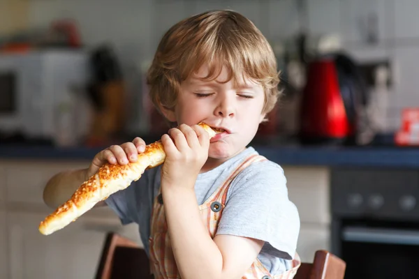 Little boy eating long loaf bread or baguette in kitchen. — Stock Photo, Image