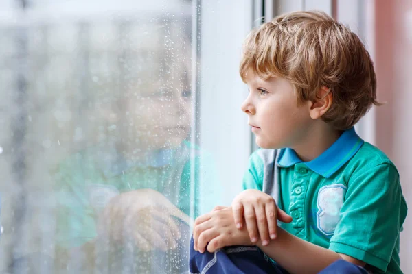 Petit garçon blond assis près de la fenêtre et regardant la goutte de pluie — Photo