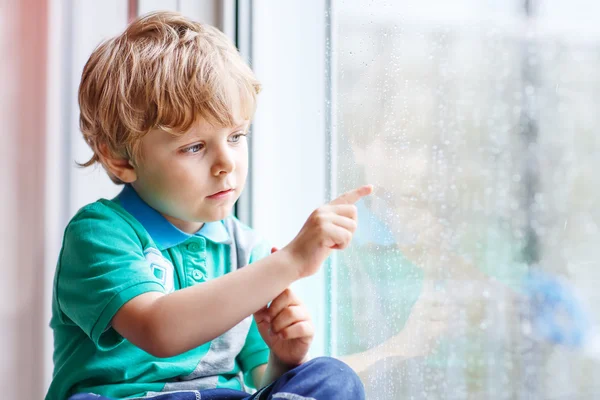 Liten blond unge pojke sitter nära fönstret och tittar på regndroppe — Stockfoto