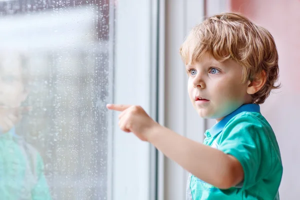 Liten blond unge pojke sitter nära fönstret och tittar på regndroppe — Stockfoto