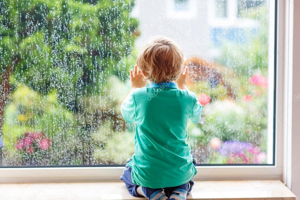 little blond kid boy sitting near window and looking on raindrop