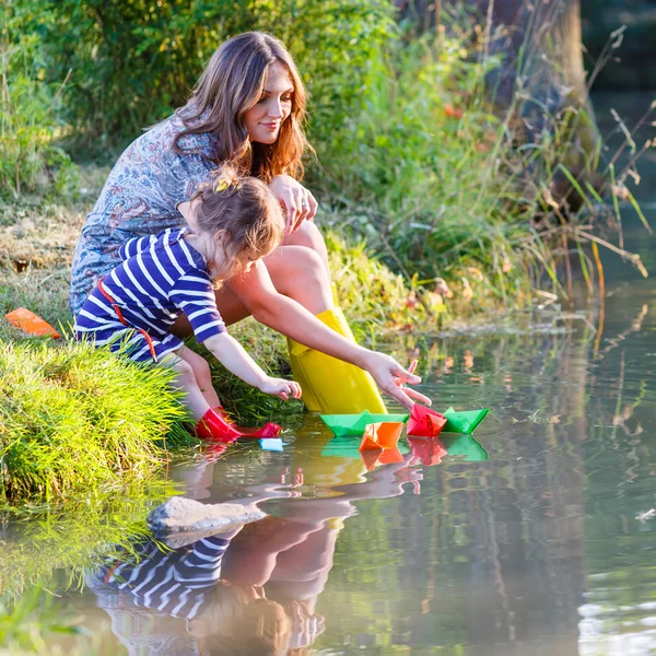 Bedårande liten flicka och hennes mamma leker med papper båtar i ett r — Stockfoto