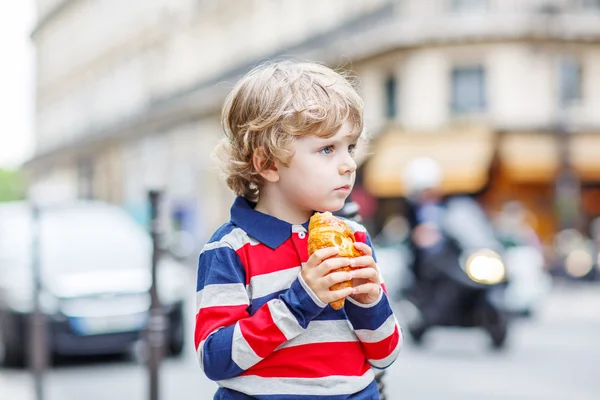 Kleines süßes Kind auf einer Straße der Stadt, das frisches Croissant isst — Stockfoto