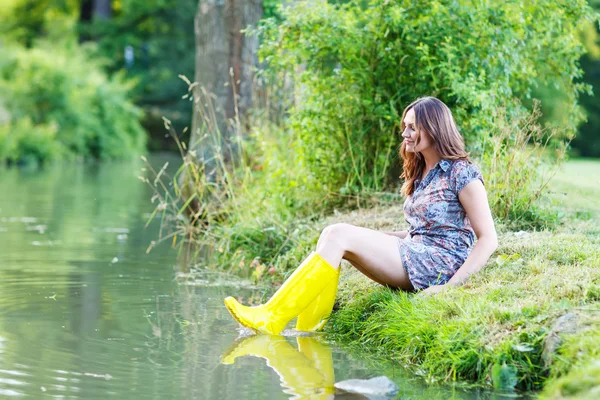 Jeune belle femme assise dans des bateaux en caoutchouc de pluie jaune par un r — Photo